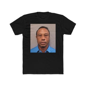 May include: Black t-shirt featuring a square image of a man in a blue collared shirt. The image appears to be a mugshot. The t-shirt is made of a soft material and has short sleeves.