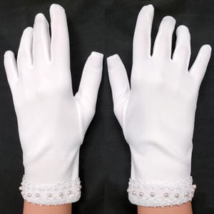 Op de afbeelding: Een paar witte handschoenen met een decoratieve kralenrand bij de pols. De rand heeft kleine parels en heldere kralen. De handschoenen zijn gemaakt van een gladde, ondoorzichtige stof.