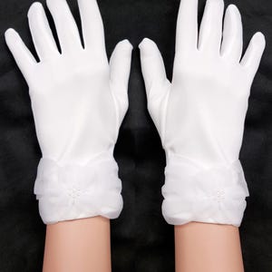 Può includere: Un paio di guanti bianchi con un design floreale sul polso. I guanti sono realizzati in un materiale liscio e opaco. Il dettaglio del polso presenta un tessuto trasparente e arruffato e un gruppo di piccoli abbellimenti rotondi.