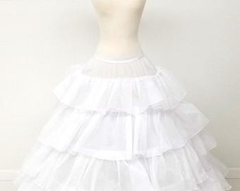 Puede incluir: Un enagua blanca de varias capas, exhibida en un maniquí. La enagua tiene múltiples capas de tela con volantes, creando una forma completa y voluminosa. El maniquí es de color neutro y el fondo es liso.
