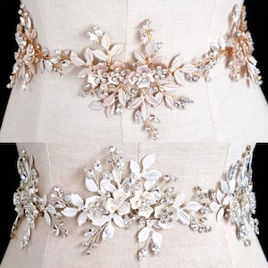 Puede incluir: Dos cinturones de novia con diseños florales y de hojas. Uno es dorado con detalles rosas y transparentes. El otro es plateado con detalles blancos y transparentes. Ambos cinturones están adornados con cristales y flores.