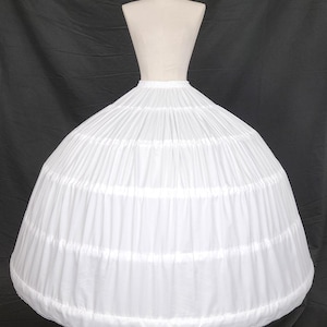 May include: A white crinoline petticoat with multiple horizontal tiers, displayed on a mannequin. The petticoat is made of a lightweight fabric, creating a full, rounded shape. The mannequin is set against a black backdrop.
