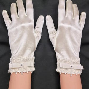 Op de afbeelding: Een paar ivoorkleurige satijnen handschoenen met delicate kanten afwerking en kleine strikdetails bij de pols. De handschoenen worden gepresenteerd tegen een donkere achtergrond, wat hun elegante ontwerp benadrukt. Deze handschoenen zijn geschikt voor formele evenementen.