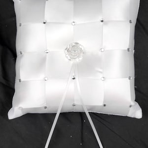 May include: White satin ring pillow with a grid pattern, adorned with small rhinestones at each intersection. A clear, decorative rose is centered, with white ribbons extending from it. A classic wedding accessory.
