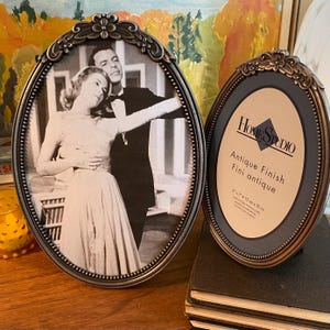May include: Two oval picture frames. One holds a black and white photo of a dancing couple. The other frame features the text "Home Studio Antique Finish" and dimensions of 13 cm x 18 cm. Both frames have decorative, dark metal borders.