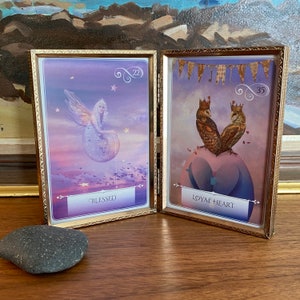 Puede incluir: Dos tarjetas con marco dorado y fondos morados. La tarjeta de la izquierda presenta un hada con alas y una corona sentada en una luna con el número 22 y la palabra "Blessed". La tarjeta de la derecha presenta dos búhos con coronas sentados en un corazón con el número 35 y las palabras "Loyal Heart".
