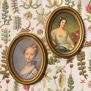 Puede incluir: Dos retratos ovalados de mujeres enmarcados en oro. El retrato de la izquierda presenta a una joven con cabello castaño claro que lleva un vestido azul y blanco. El retrato de la derecha presenta a una mujer con cabello castaño que lleva un vestido blanco con ribete dorado y un pájaro marrón en el hombro.