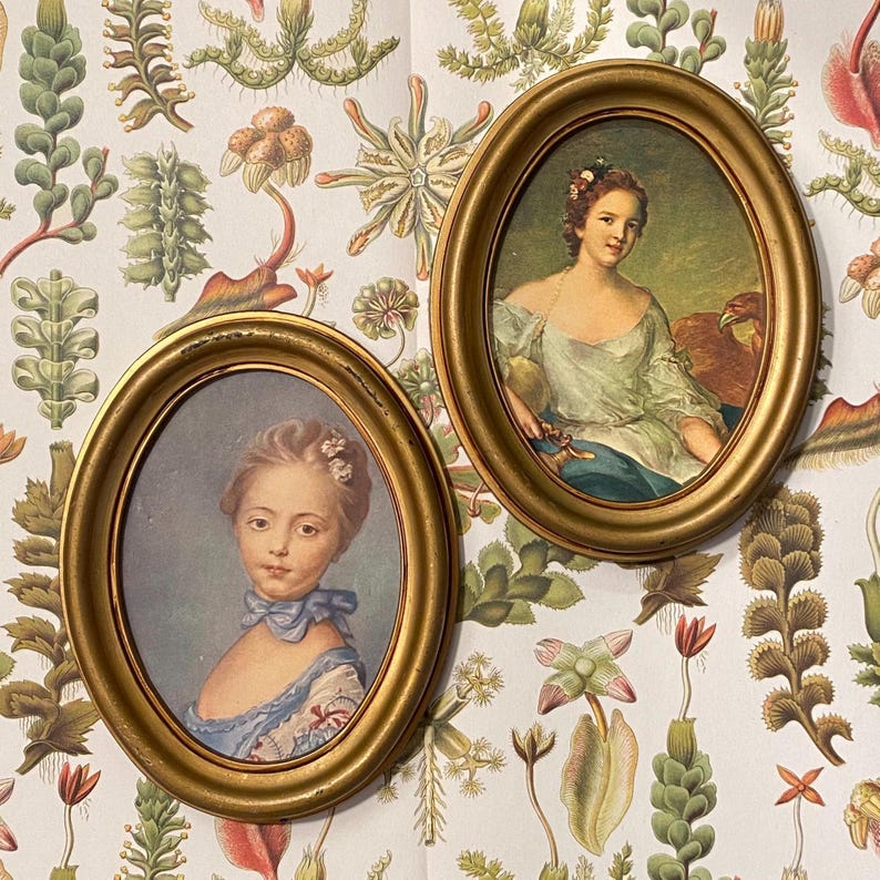 May include: Two oval gold framed portraits of women. The portrait on the left features a young woman with light brown hair wearing a blue and white dress. The portrait on the right features a woman with brown hair wearing a white dress with a gold trim and a brown bird on her shoulder.