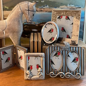 Peut inclure: Une collection de cadres photo argentés avec un motif de rouge-gorge. Les cadres sont disposés sur une surface en bois avec une figurine de cheval en bois en arrière-plan.