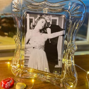 Könnte beinhalten: Ein klarer, verzierter Glasbilderrahmen mit einem Schwarz-Weiß-Foto eines Paares. Der Rahmen hat einen dekorativen, gewellten Rand. Einige kleine, herzförmige Pralinen und Lichterketten befinden sich im Vordergrund.