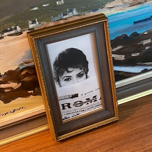 Puede incluir: Un marco de fotos rectangular con acabado dorado metálico y un borde interior gris oscuro. El marco contiene una fotografía en blanco y negro de una persona con cabello oscuro sosteniendo un periódico con la palabra ROMA visible.