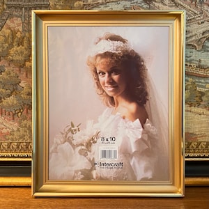 May include: Gold-colored picture frame with a white mat and a photo of a bride wearing a white dress and veil. The frame is labeled "8 x 10" and "20 x 25 cm" and has the Intercraft logo with the text "The Frame People".