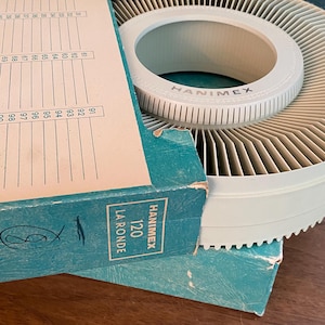 May include: A vintage Hanimex 120 slide carousel and storage box. The carousel is light green with a circular design and the brand name printed on the side. The box is teal with white text and a numbered chart.