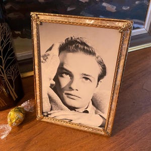 May include: A vintage framed black and white portrait of a man. The gold frame has a decorative border. The portrait is on a wooden surface, with a decorative vase and wrapped sweet in the background.