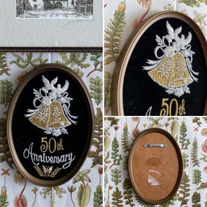 May include: A gold-framed embroidery hoop with a black velvet background. The embroidery depicts two white doves with gold bells and the text "50th Anniversary".