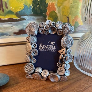 Puede incluir: Un marco de fotos de peltre plateado decorado con varios botones y una concha. El marco tiene un fondo azul con el texto "Seagull Pewter".