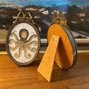 May include: Two oval-shaped brass picture frames with a decorative bow on top. One frame has a vintage illustration of an octopus inside. The other frame has a brown backing.