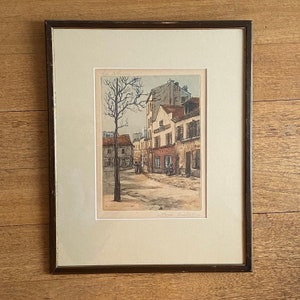 May include: A framed black and white print of a street scene in Paris, France. The print depicts a narrow street with buildings on either side, a tree in the centre, and people walking on the pavement. The print is titled "Rue de la Sorbonne" and signed "Paul Jouve".