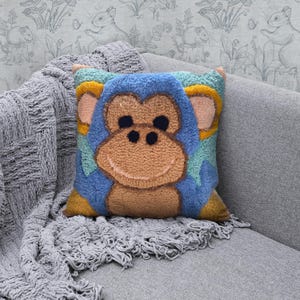 Può includere: Un cuscino di peluche blu e giallo con un viso di scimmia sorridente al centro. Gli occhi della scimmia sono neri e il suo naso è marrone. Il cuscino è su un divano grigio.