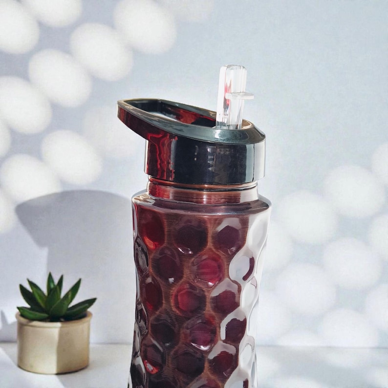 May include: A clear water bottle with a textured, honeycomb-like pattern. The bottle is filled with a dark red liquid and has a black lid with a clear straw. A small succulent plant in a beige pot is in the background.
