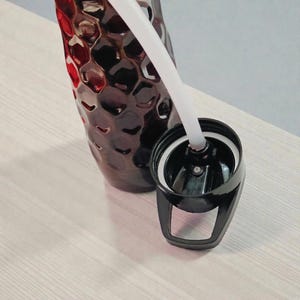 May include: A dark red water bottle with a textured surface and a black lid. A white straw extends from the lid. The bottle is partially visible, resting on a light-colored surface.