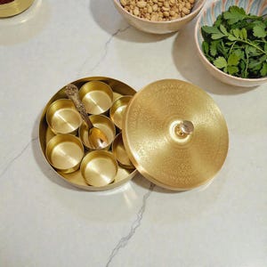 Könnte beinhalten: Ein runder, goldfarbener Gewürzbehälter mit dekorativem Deckel und kleinem Löffel. Der Behälter ist geöffnet und zeigt mehrere kleine Fächer. Der Deckel hat ein verziertes Muster. Schalen mit Zutaten befinden sich im Hintergrund.