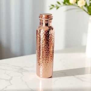 Puede incluir: Una botella de agua de cobre con textura martillada. La botella tiene una tapa de rosca y forma cilíndrica. El cobre tiene un tono marrón rojizo cálido. La botella está sobre una superficie de mármol blanco.