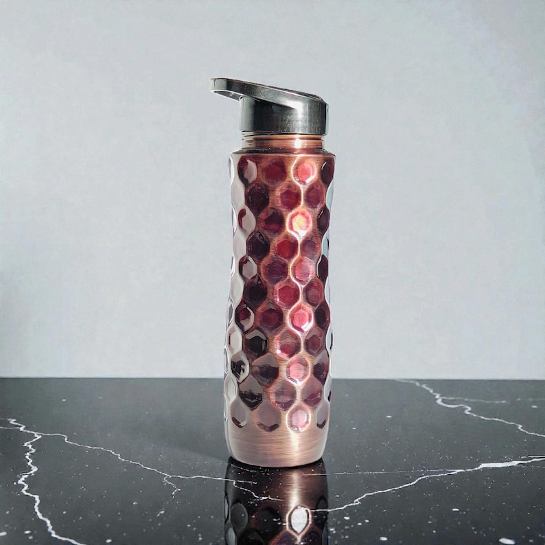 May include: A copper-colored water bottle with a textured, honeycomb-like pattern. The bottle has a silver-colored lid with a flip-top opening. The base of the bottle is a darker copper color. The bottle is designed for hydration.