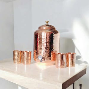 May include: A copper water dispenser with a hammered texture, a spigot, and a lid with a gold-colored finial. It is accompanied by six matching copper cups, all arranged on a light-colored surface.