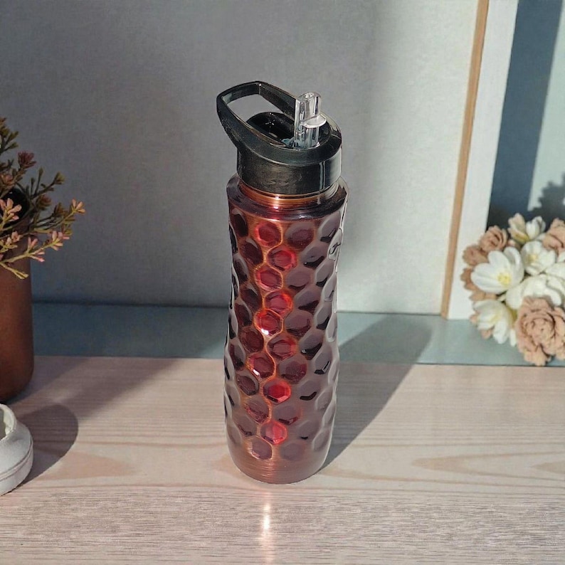 May include: A red and bronze water bottle with a black lid and a built-in straw. The bottle has a textured, honeycomb-like pattern. The bottle is sitting on a light-colored wooden surface.