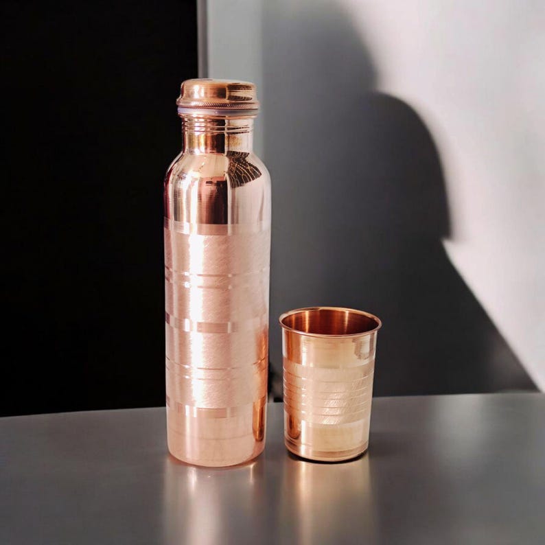May include: A copper water bottle and a matching cup. The bottle has a screw-on lid and etched horizontal lines. The cup is cylindrical with similar etched lines. Both items have a shiny, metallic finish and are displayed on a reflective surface.