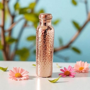 Pu&ograve; includere: Una borraccia in rame color oro rosa con una texture martellata e un tappo a vite. La borraccia &egrave; circondata da margherite rosa e foglie verdi su uno sfondo azzurro.