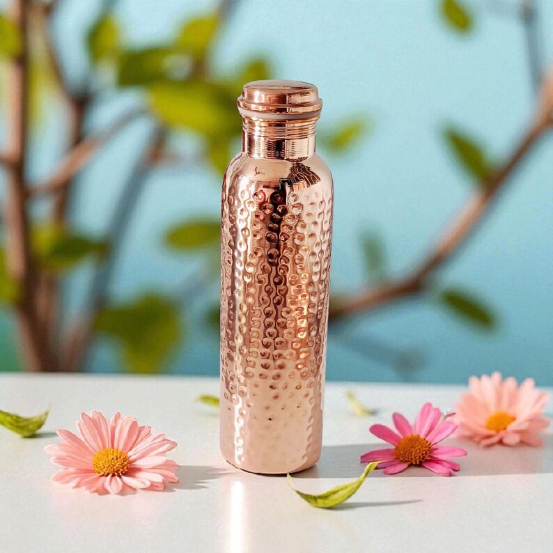 Pu&ograve; includere: Una borraccia in rame color oro rosa con una texture martellata e un tappo a vite. La borraccia &egrave; circondata da margherite rosa e foglie verdi su uno sfondo azzurro.