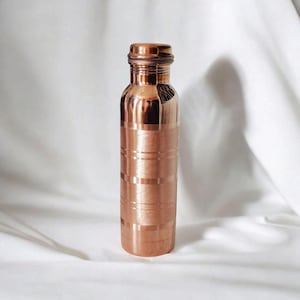 May include: A copper water bottle with a screw-on cap. The bottle has horizontal etched lines around the body. The copper has a shiny, reflective surface. The bottle is cylindrical and stands upright against a white background.