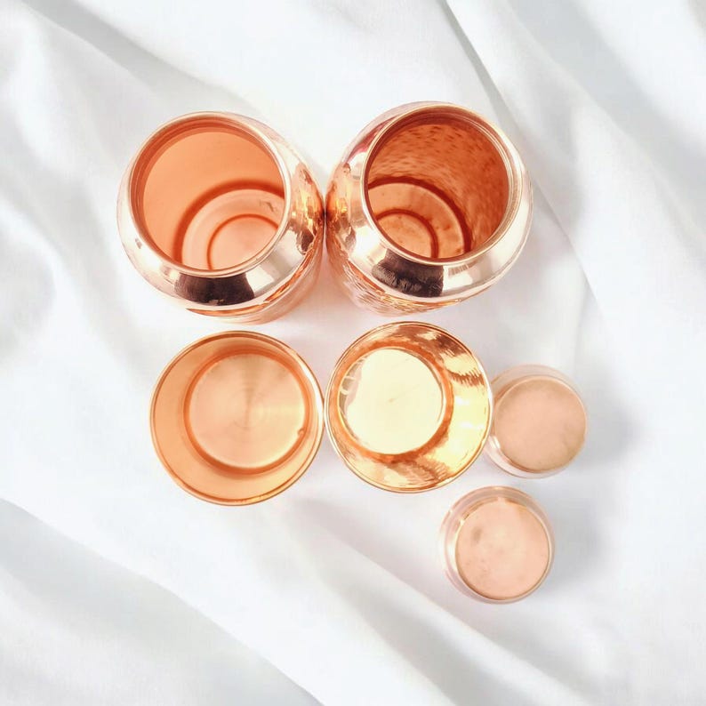 May include: An assortment of copper-toned containers and lids displayed on a white background. The arrangement features two large, open-topped vessels, two lids, and three smaller containers with lids. The metallic finish reflects light, giving a warm, inviting appearance.