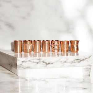 May include: Seven copper-coloured drinking glasses with different textures and designs are displayed in a row. The glasses sit on a white marble surface, highlighting their unique patterns and shapes. The glasses are made of copper and have a shiny finish.