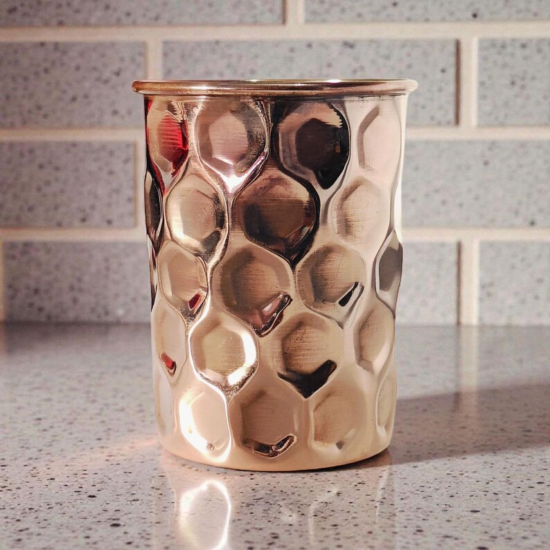 May include: A copper-colored, hammered metal cup with a cylindrical shape. The cup has a textured surface with a pattern of rounded, irregular shapes. The rim of the cup is smooth and polished. The cup is likely used for drinking or as a decorative item.