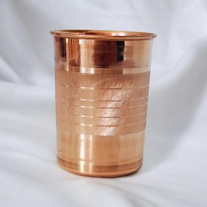 May include: A copper-coloured drinking glass with a cylindrical form. The glass features horizontal etched lines around the mid-section, providing texture. The rim and base are smooth, reflecting light. The design is simple and elegant.