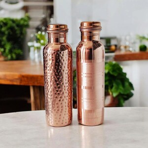 May include: Two copper water bottles with screw-on lids. One bottle has a hammered texture, and the other has horizontal stripes. Both bottles are a shiny, warm copper colour and sit on a white surface. The background is blurred, with hints of greenery.