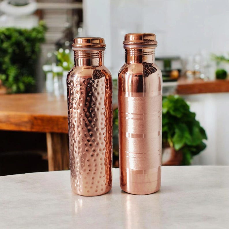 May include: Two copper water bottles with screw-on lids. One bottle has a hammered texture, and the other has horizontal stripes. Both bottles are a shiny, warm copper colour and sit on a white surface. The background is blurred, with hints of greenery.
