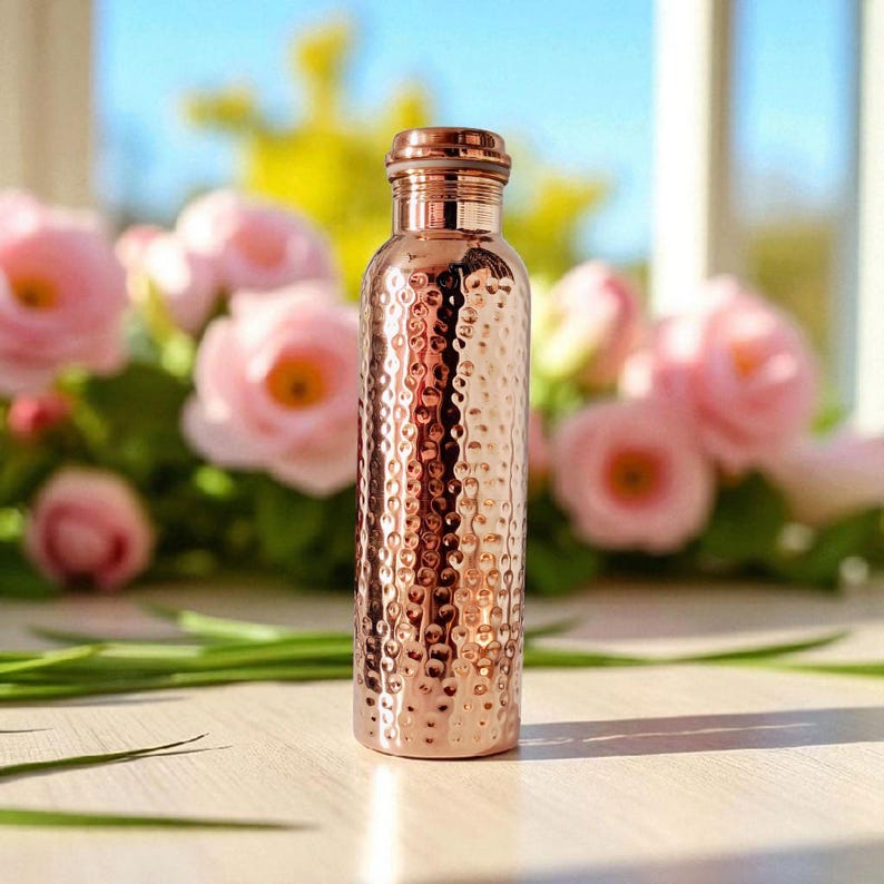 Pu&ograve; includere: Una borraccia color rame con una texture martellata. La bottiglia ha una parte superiore arrotondata e un tappo abbinato. Lo sfondo presenta fiori rosa e verde, con una vista sfocata di una finestra e cielo blu.
