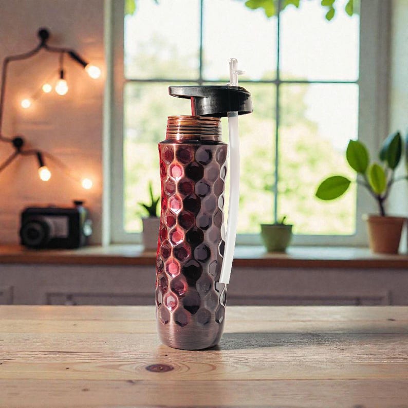 May include: A stainless steel water bottle with a textured, honeycomb-like pattern in shades of red and silver. It has a black lid with a white straw. The bottle is on a wooden surface, with a window and plants in the background.