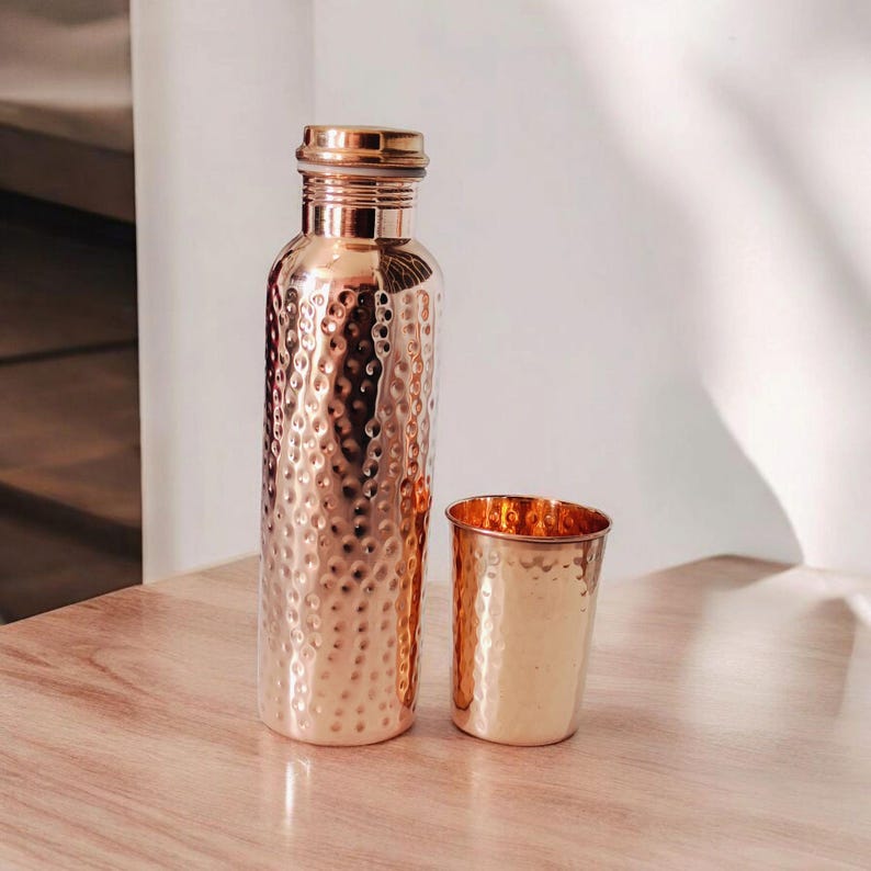 May include: A copper water bottle and a matching cup are displayed on a wooden surface. The bottle features a hammered texture and a screw-on lid. The cup, also with a hammered texture, is a smaller version of the bottle. Both items are a warm copper colour.
