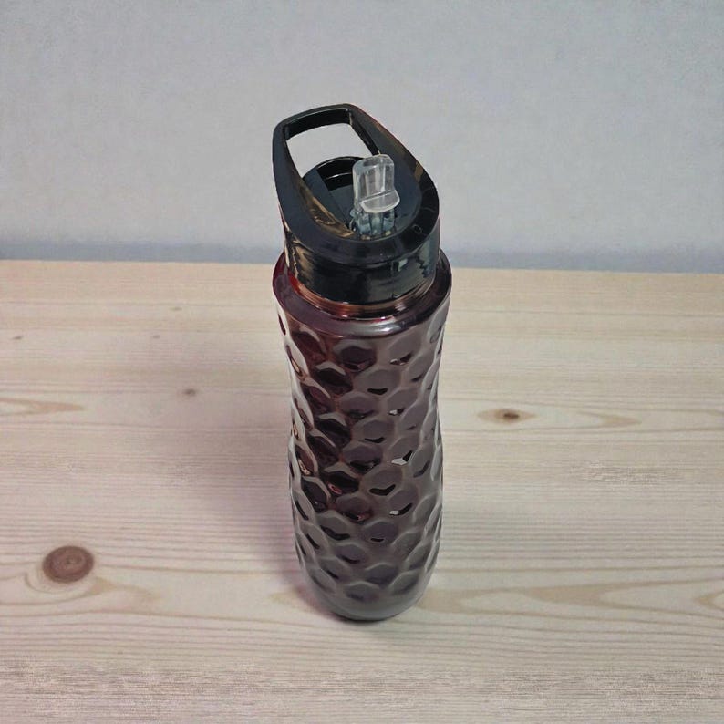 May include: A maroon water bottle with a black lid and handle. The bottle has a textured, honeycomb-like pattern. The lid features a clear, flip-top spout. The bottle is designed for hydration and is suitable for various activities.