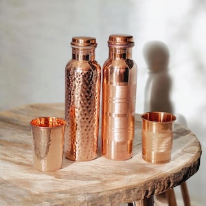May include: Two copper water bottles and two copper cups are displayed on a wooden table. One bottle has a hammered texture, the other has horizontal lines. The cups are copper, one with a hammered texture, the other with horizontal lines. A decorative set.