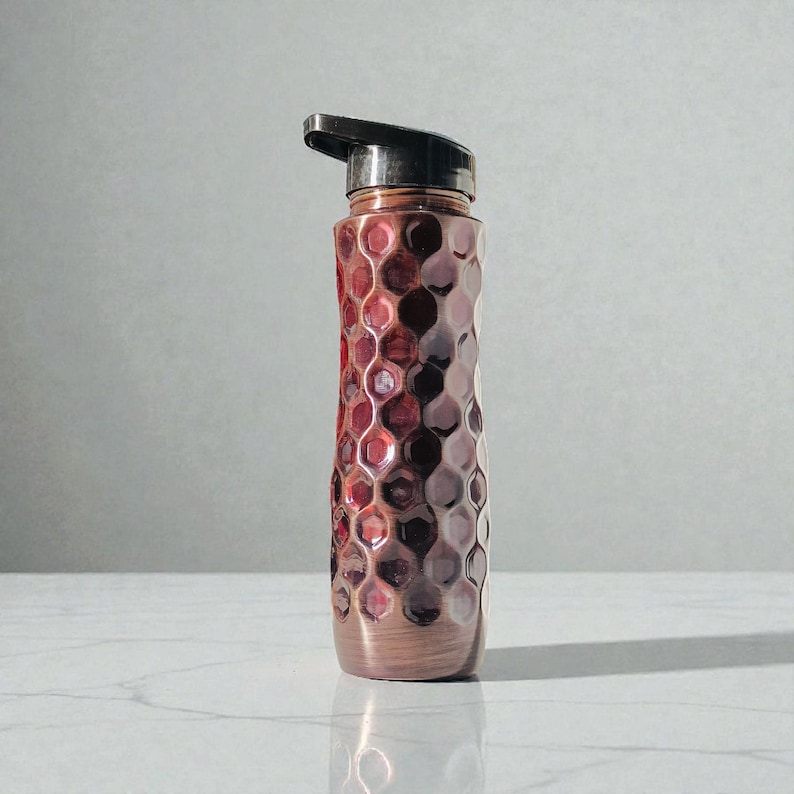May include: A copper-colored water bottle with a textured, honeycomb-like pattern. The bottle has a black flip-top lid and a metallic finish. The design features shades of red and brown, creating a visually appealing and functional item.