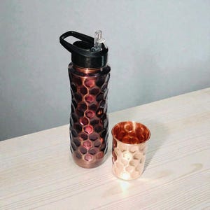 May include: A copper-colored water bottle with a black lid and a matching copper-colored cup. The bottle has a textured, honeycomb-like pattern. The cup has a similar hammered design. Both items are on a light-colored wooden surface.