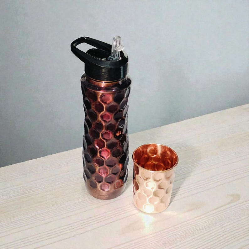May include: A copper-colored water bottle with a black lid and a matching copper-colored cup. The bottle has a textured, honeycomb-like pattern. The cup has a similar hammered design. Both items are on a light-colored wooden surface.