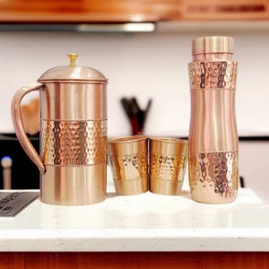 May include: A copper pitcher with a handle and lid, two matching copper tumblers, and a tall copper water bottle. Each item features a polished finish with a central band of hammered texture and small beaded detailing along the edges of the band.
