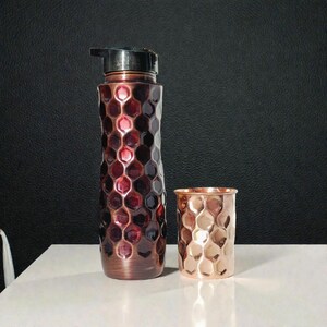 May include: A copper-colored water bottle and a matching tumbler. The bottle has a black cap and a honeycomb pattern. The tumbler is a smaller version of the bottle, also with a honeycomb design. Both items are on a white surface.